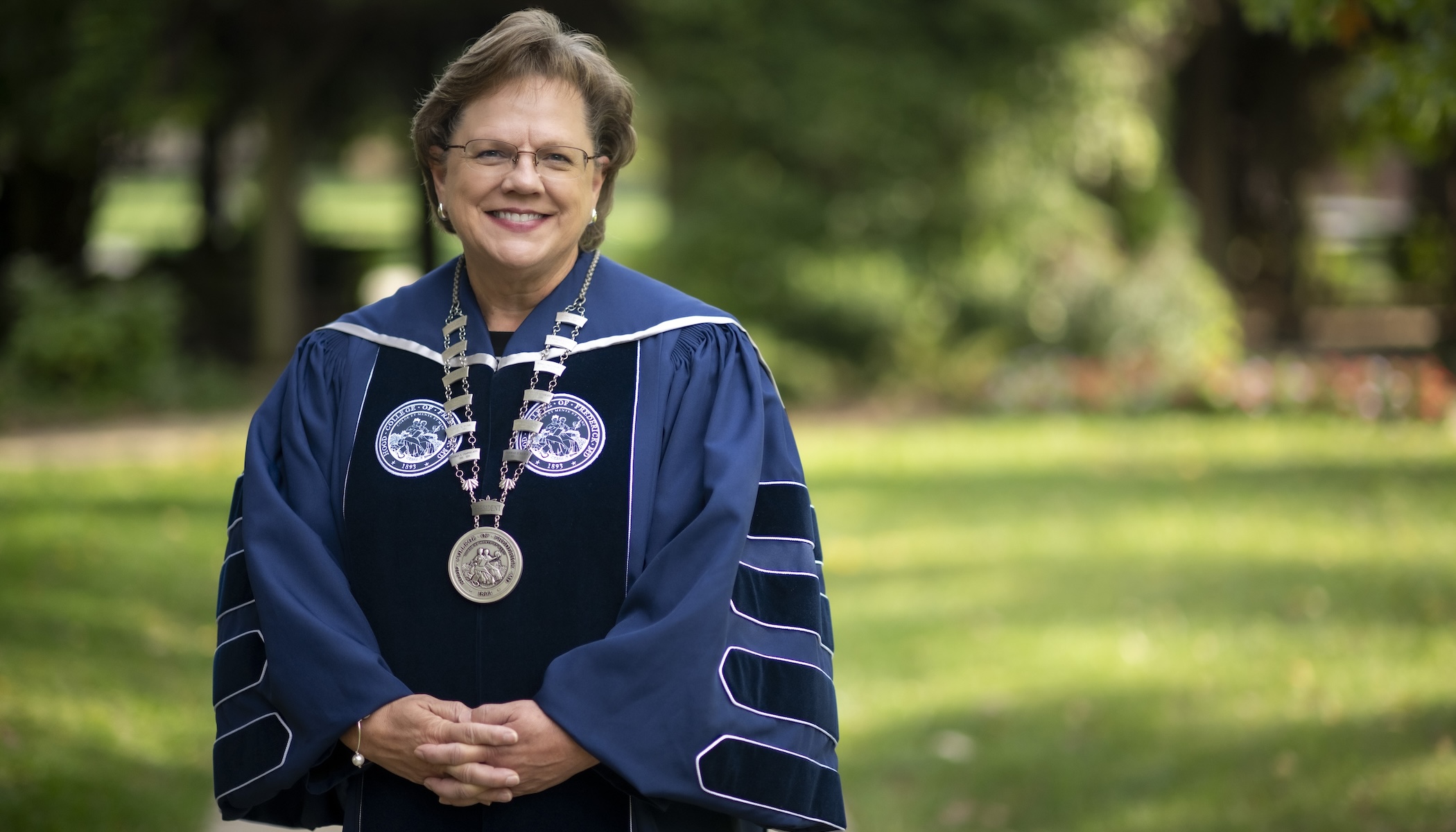 A headshot of Hood College President Debbie Ricker, Ph.D., wearing presidential regailia outside in front of a lush, green lawn