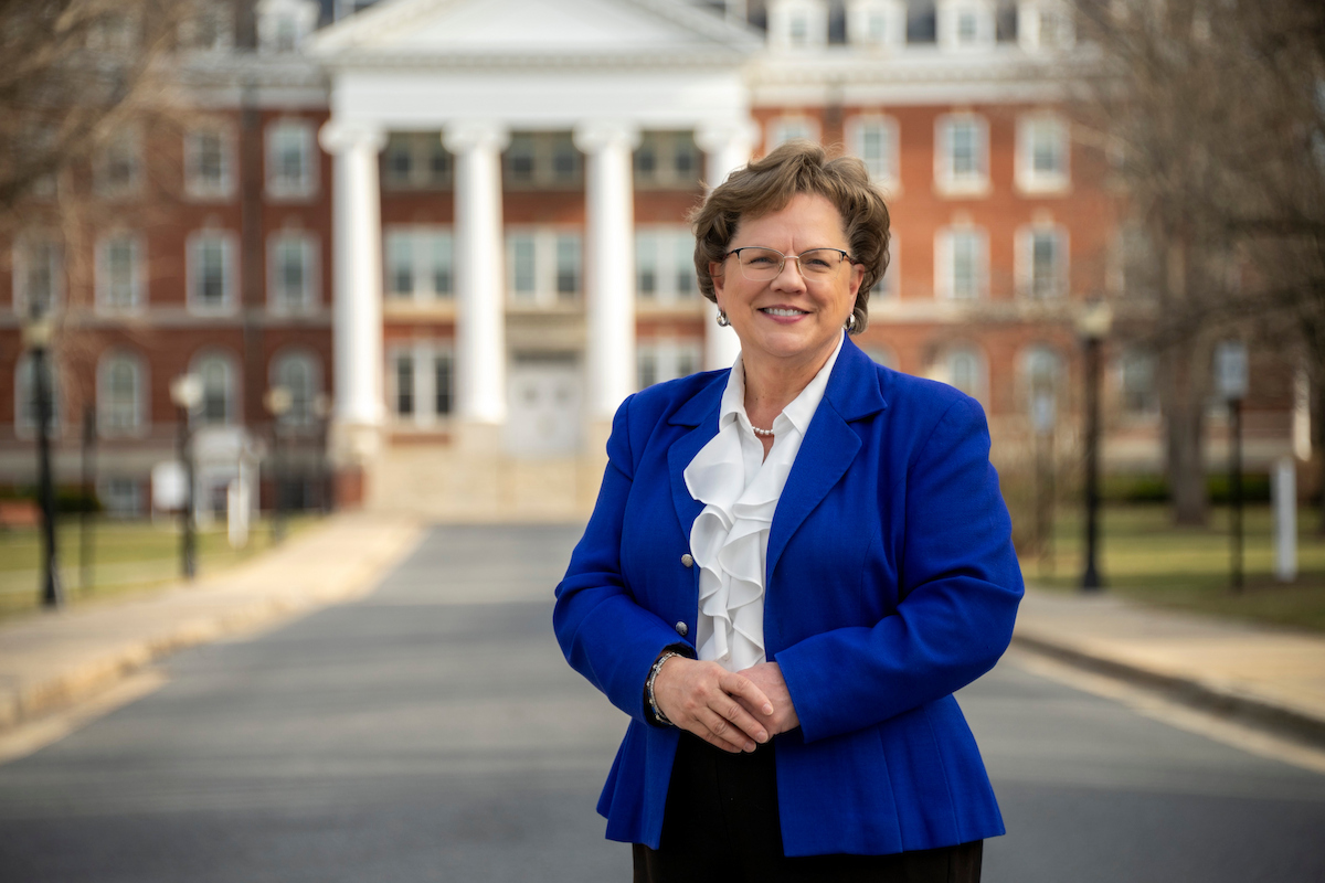 President Debbie Ricker, wearing a blue blazer and frilly white shirt, stands in the street in front of four large pillars