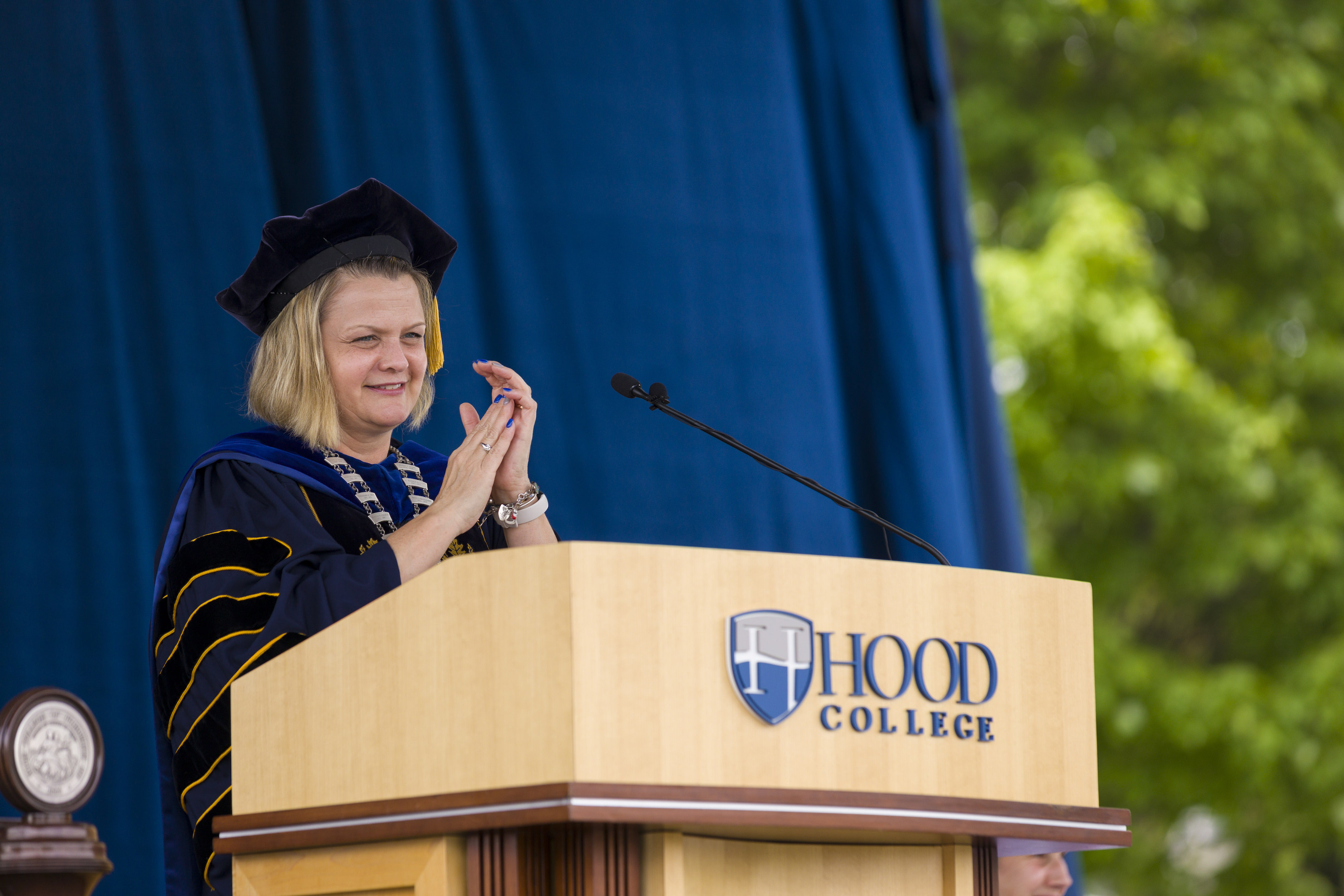 Hood College President Andrea E. Chapdelaine claps as she issues remarks from the podium at the class of 2021 graduation ceremony