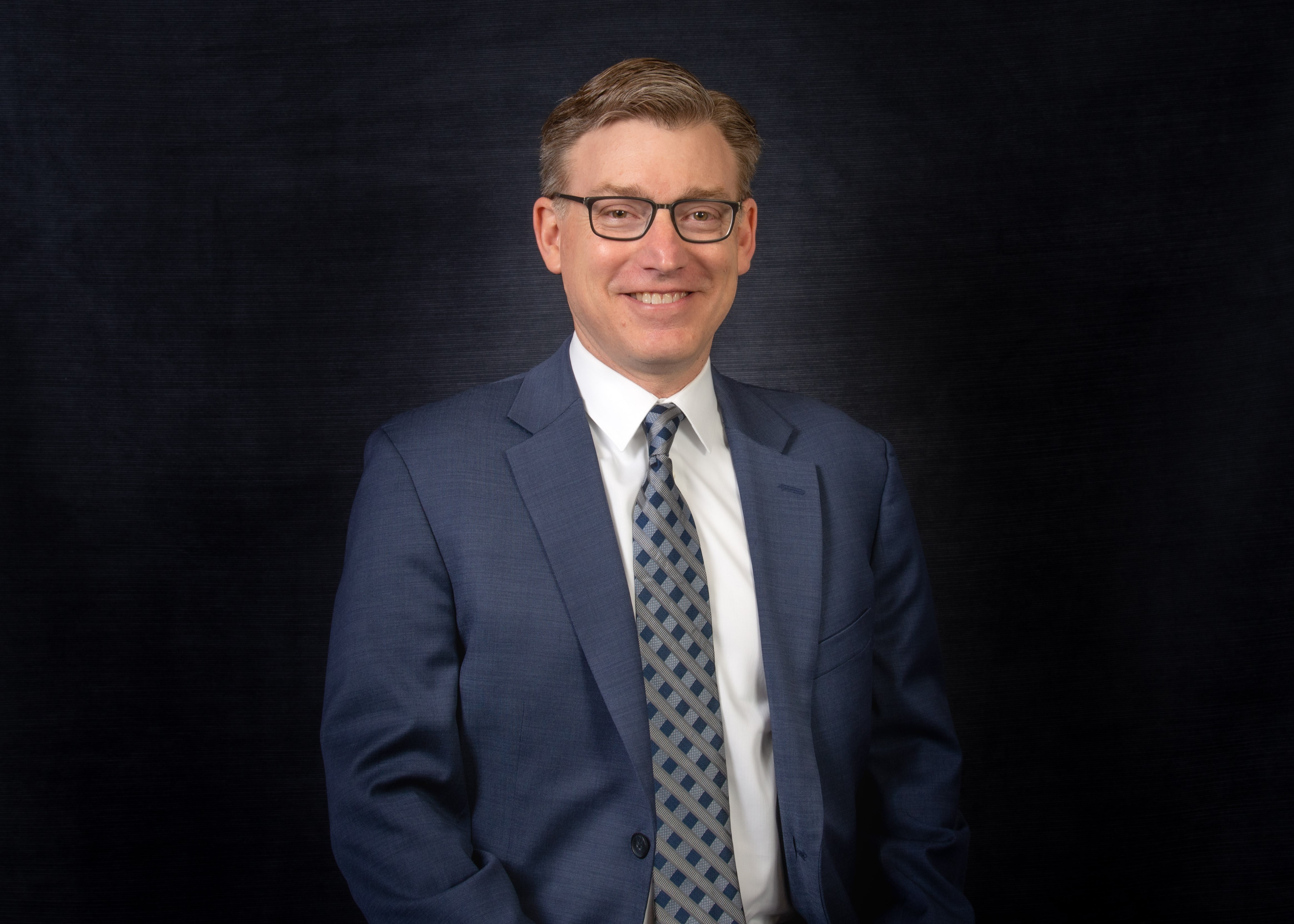 A headshot of a smiling robert klinedinst, taken in front of a dark background