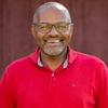 Headshot of Haroun Hallack. He is wearing a vibrant red shirt and glasses in front of a red wooden building.